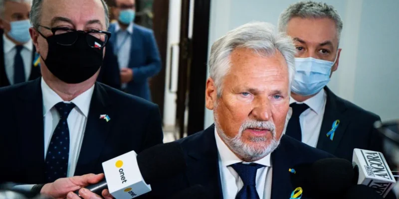 Na zdjęciu widoczni są mężczyźni w garniturach, w tym jeden z siwą brodą, mówiący do mikrofonów, podczas konferencji prasowej.