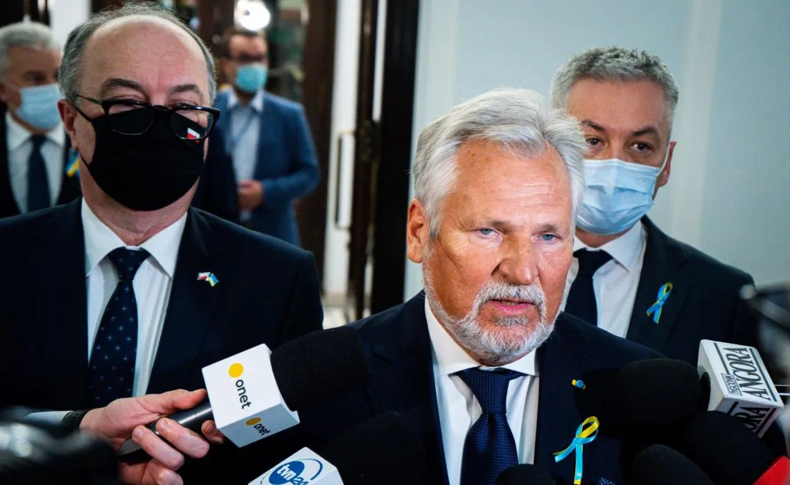 Na zdjęciu widoczni są mężczyźni w garniturach, w tym jeden z siwą brodą, mówiący do mikrofonów, podczas konferencji prasowej.