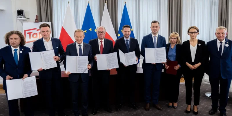 Na zdjęciu grupa dziewięciu osób stoi przy stole, trzymając dokumenty, w tle widoczne flagi Polski i Unii Europejskiej.