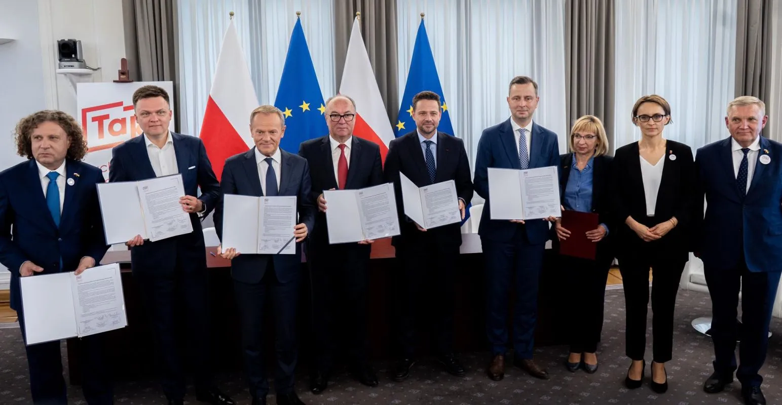 Na zdjęciu grupa dziewięciu osób stoi przy stole, trzymając dokumenty, w tle widoczne flagi Polski i Unii Europejskiej.