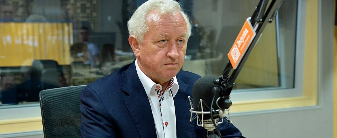 Starszy mężczyzna w eleganckiej marynarce siedzi w studiu radiowym i mówi do mikrofonu.