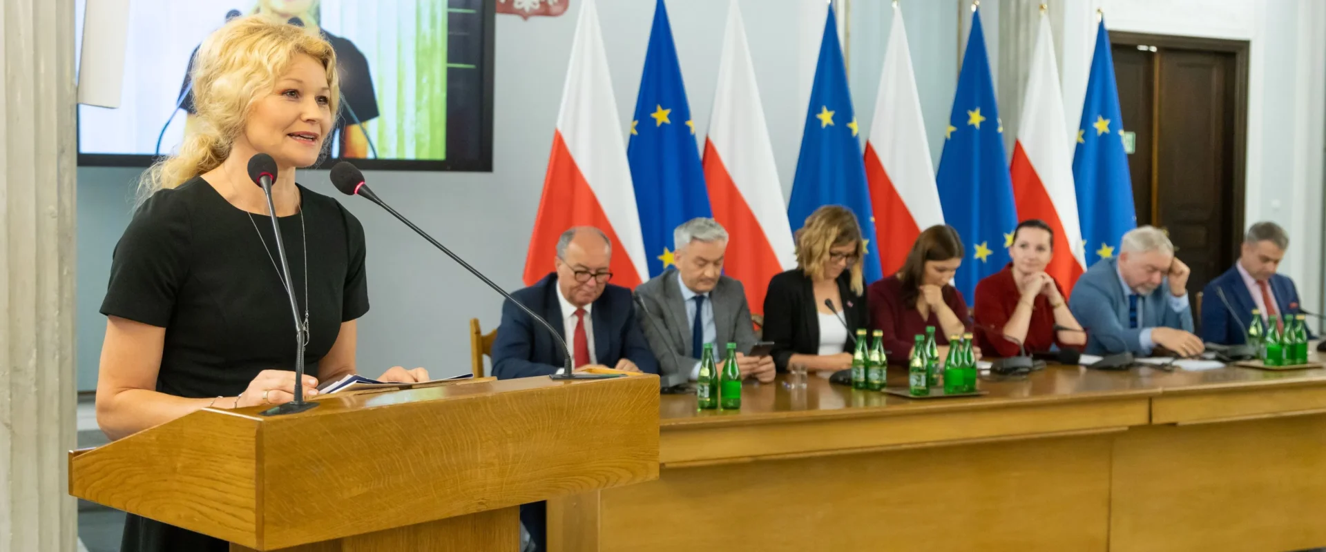 Kobieta przemawia z mównicy podczas konferencji samorządowców Lewicy; za nią siedzi grupa urzędników przy stole, a w tle widać flagi Polski i Unii Europejskiej oraz duży ekran z jej wizerunkiem.