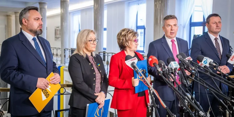 Na zdjęciu pięciu polityków stojących przy mównicy otoczonej mikrofonami, wygłaszających przemówienie w jasnym wnętrzu.