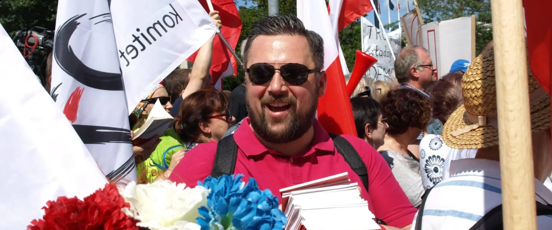 Uśmiechnięty mężczyzna w czerwonej koszulce i ciemnych okularach trzyma stos broszur w tłumie demonstrantów z biało‑czerwonymi flagami, a na pierwszym planie widać wianek z kolorowych kwiatów.