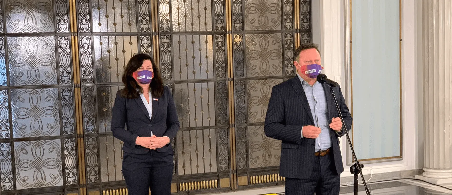 Dwoje osób w formalnych garniturach stoi w holu z ozdobnymi metalowymi drzwiami; po prawej stronie mężczyzna mówi do mikrofonu ustawionego na statywie, po lewej stronie stoi kobieta.