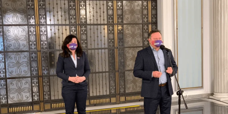 Dwoje osób w formalnych garniturach stoi w holu z ozdobnymi metalowymi drzwiami; po prawej stronie mężczyzna mówi do mikrofonu ustawionego na statywie, po lewej stronie stoi kobieta.