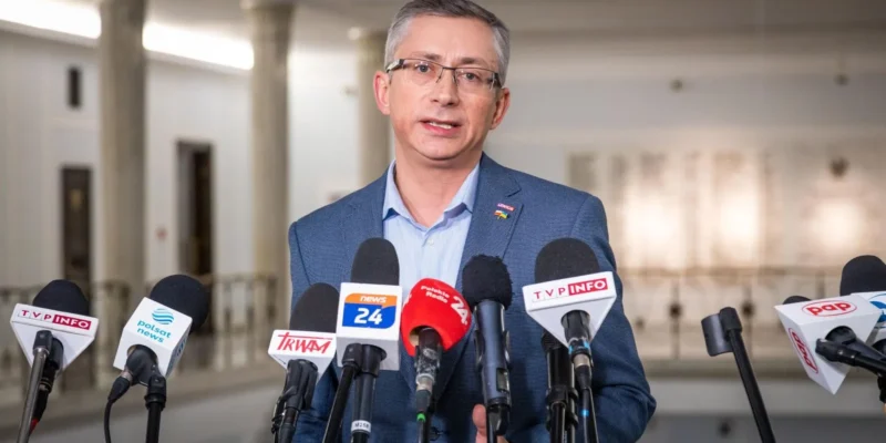 Mężczyzna w niebieskim garniturze i okularach przemawia do licznych mikrofonów dziennikarskich podczas konferencji prasowej.