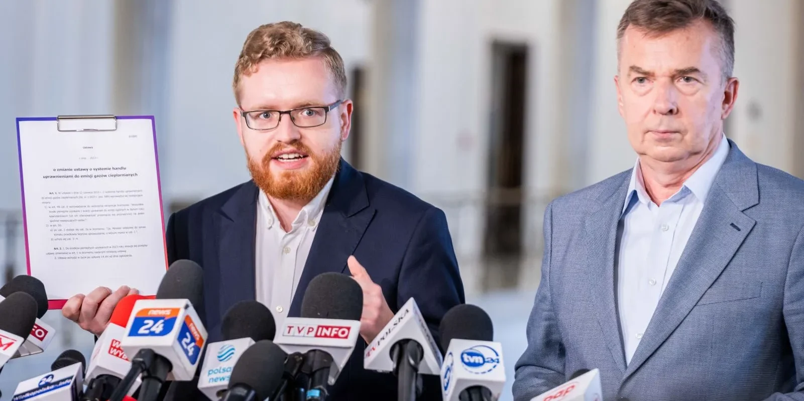 Dwóch mężczyzn w garniturach podczas konferencji prasowej z licznymi mikrofonami; jeden z nich trzyma dokument dotyczący 100 procent środków z ETS na modernizację energetyki i hasła „Aby Polki i Polacy mieli tańszy prąd”.
