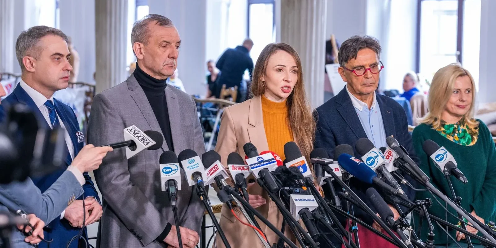 Na zdjęciu grupa ludzi stoi przed mikrofonami, prowadząc konferencję prasową w jasnym wnętrzu budynku.