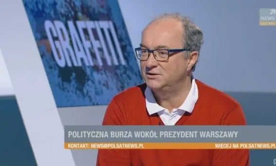 Mężczyzna w okularach i czerwonym swetrze wypowiada się w studiu telewizyjnym, na ekranie widoczny napis informujący o politycznej burzy wokół prezydenta Warszawy.
