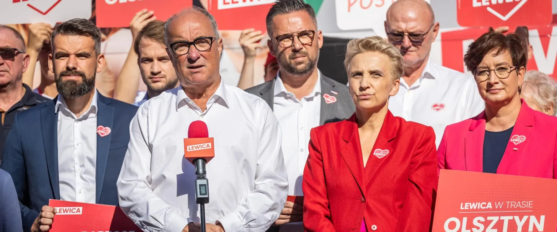 Grupa polityków Lewicy na konferencji prasowej, w centrum mężczyzna w białej koszuli przy mikrofonie, otoczona plakatami i banerami Lewicy z hasłami i nazwami miast.