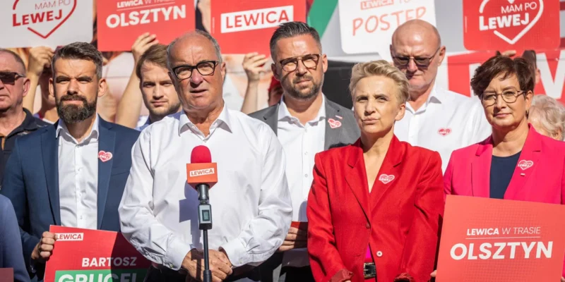 Grupa polityków Lewicy na konferencji prasowej, w centrum mężczyzna w białej koszuli przy mikrofonie, otoczona plakatami i banerami Lewicy z hasłami i nazwami miast.