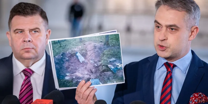 Na zdjęciu widać dwóch mężczyzn stojących przed mikrofonami, z jednym z nich trzymającym w ręku wydruk przedstawiający miejsce, które wygląda na teren z wrakiem lub zniszczonym obiektem.