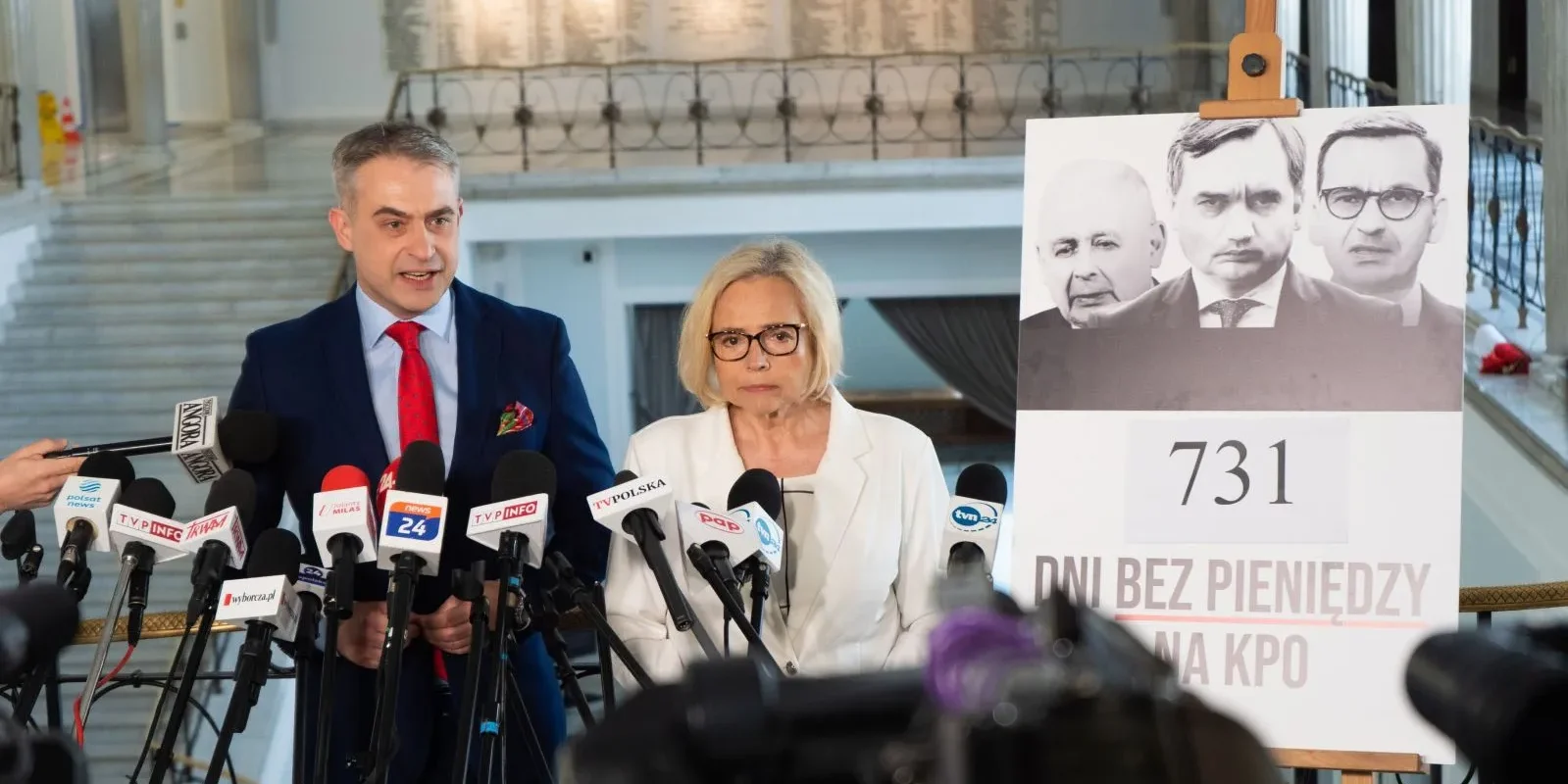 Dwóch polityków na konferencji prasowej z licznymi mikrofonami, obok nich plakat z numerem 731 i czarno-białymi portretami trzech osób.