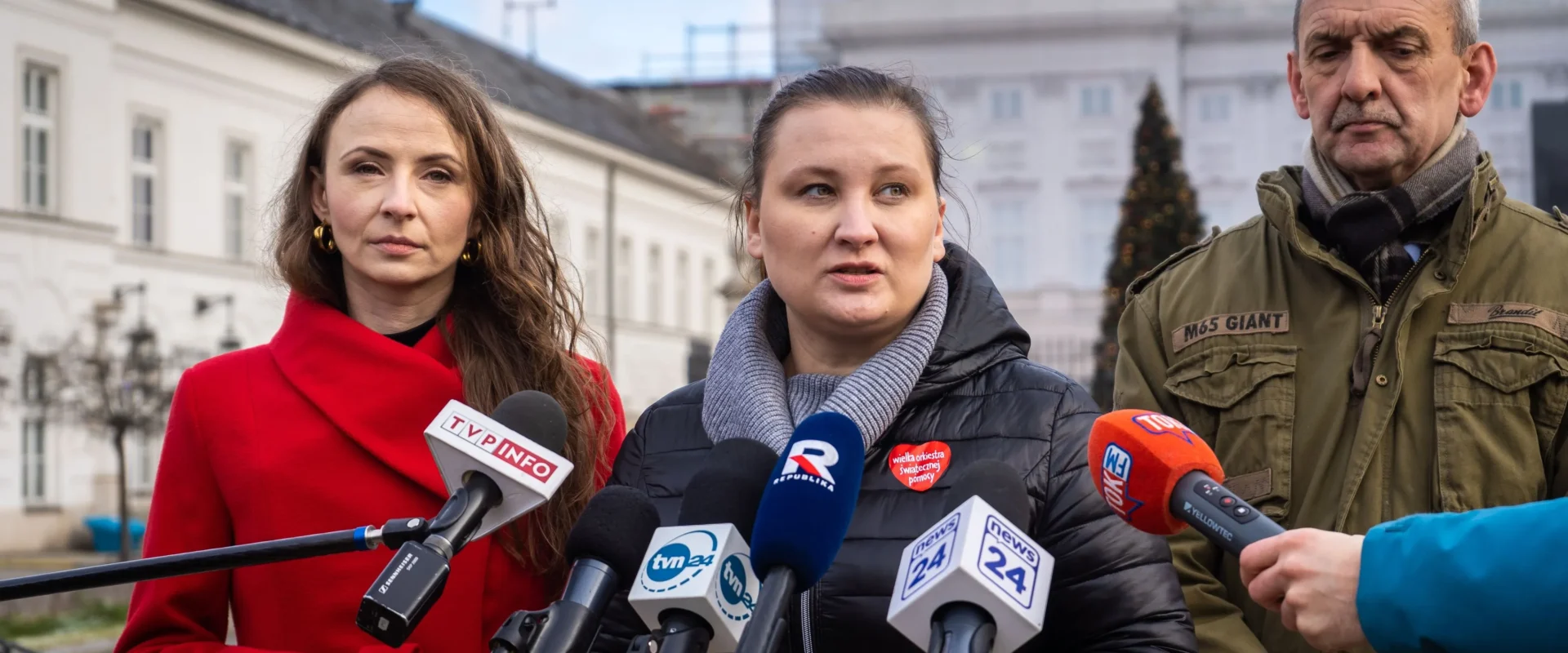 Na zdjęciu widać trzech ludzi stojących przed mikrofonami, w tle budynek oraz choinka, podczas konferencji prasowej.