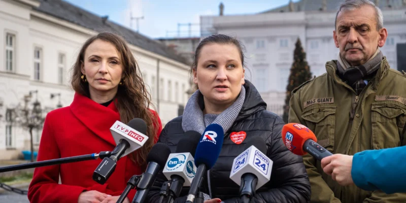 Na zdjęciu widać trzech ludzi stojących przed mikrofonami, w tle budynek oraz choinka, podczas konferencji prasowej.