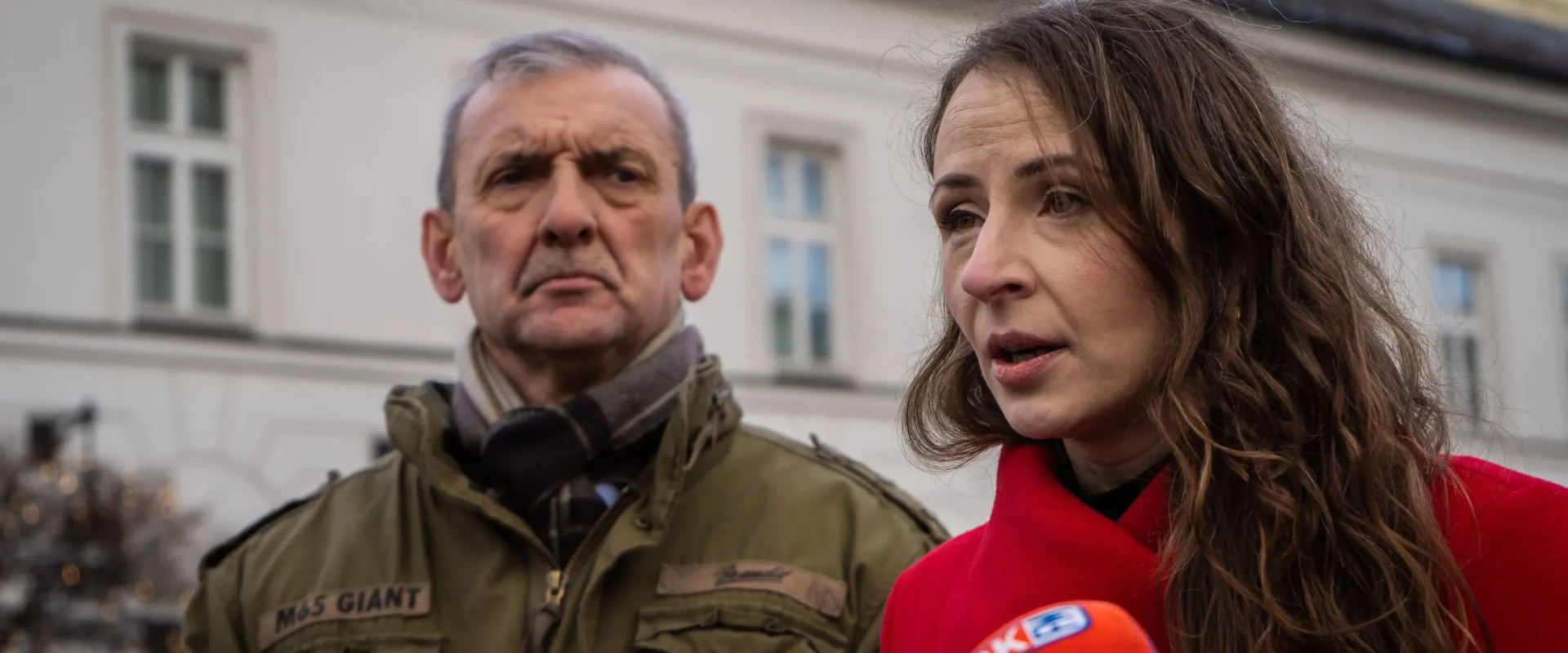 Para dorosłych udziela wywiadu przed kamerami dziennikarzy, mężczyzna w zielonej kurtce z naszywką i szalikiem oraz kobieta w czerwonym płaszu, na tle białego budynku.