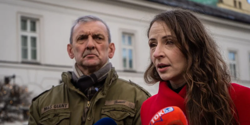 Para dorosłych udziela wywiadu przed kamerami dziennikarzy, mężczyzna w zielonej kurtce z naszywką i szalikiem oraz kobieta w czerwonym płaszu, na tle białego budynku.