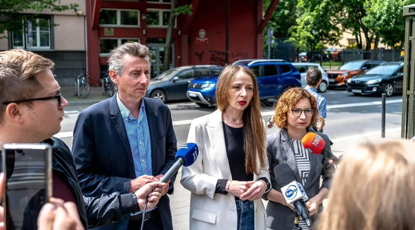 Grupa osób stoi na ulicy podczas konferencji prasowej, trzymając mikrofony z logotypami mediów.