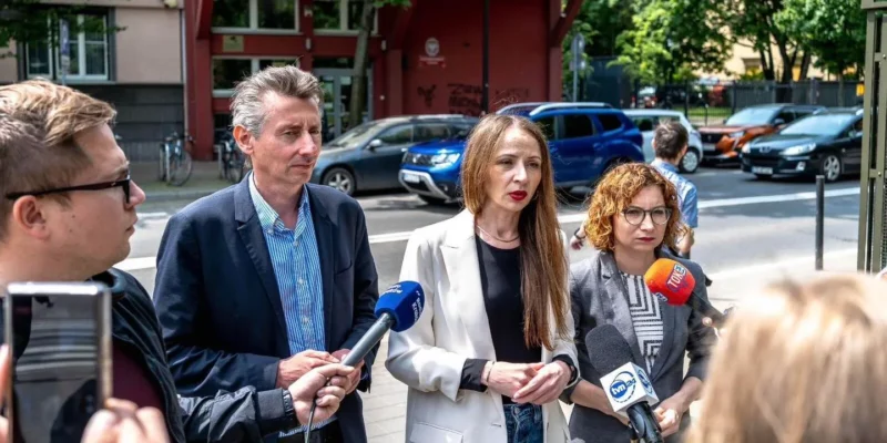 Grupa osób stoi na ulicy podczas konferencji prasowej, trzymając mikrofony z logotypami mediów.