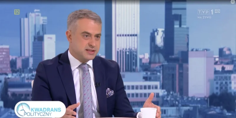 Mężczyzna w garniturze rozmawia w programie telewizyjnym przy biurku, z panoramą miasta w tle.