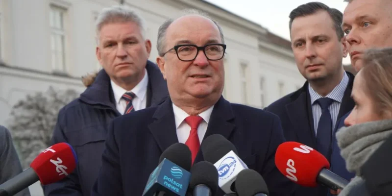 Na zdjęciu widoczny jest mężczyzna w okularach, który przemawia do zgromadzonych dziennikarzy, a w tle stoją inni mężczyźni.