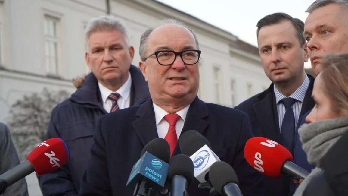 Na zdjęciu widoczny jest mężczyzna w okularach, który przemawia do zgromadzonych dziennikarzy, a w tle stoją inni mężczyźni.