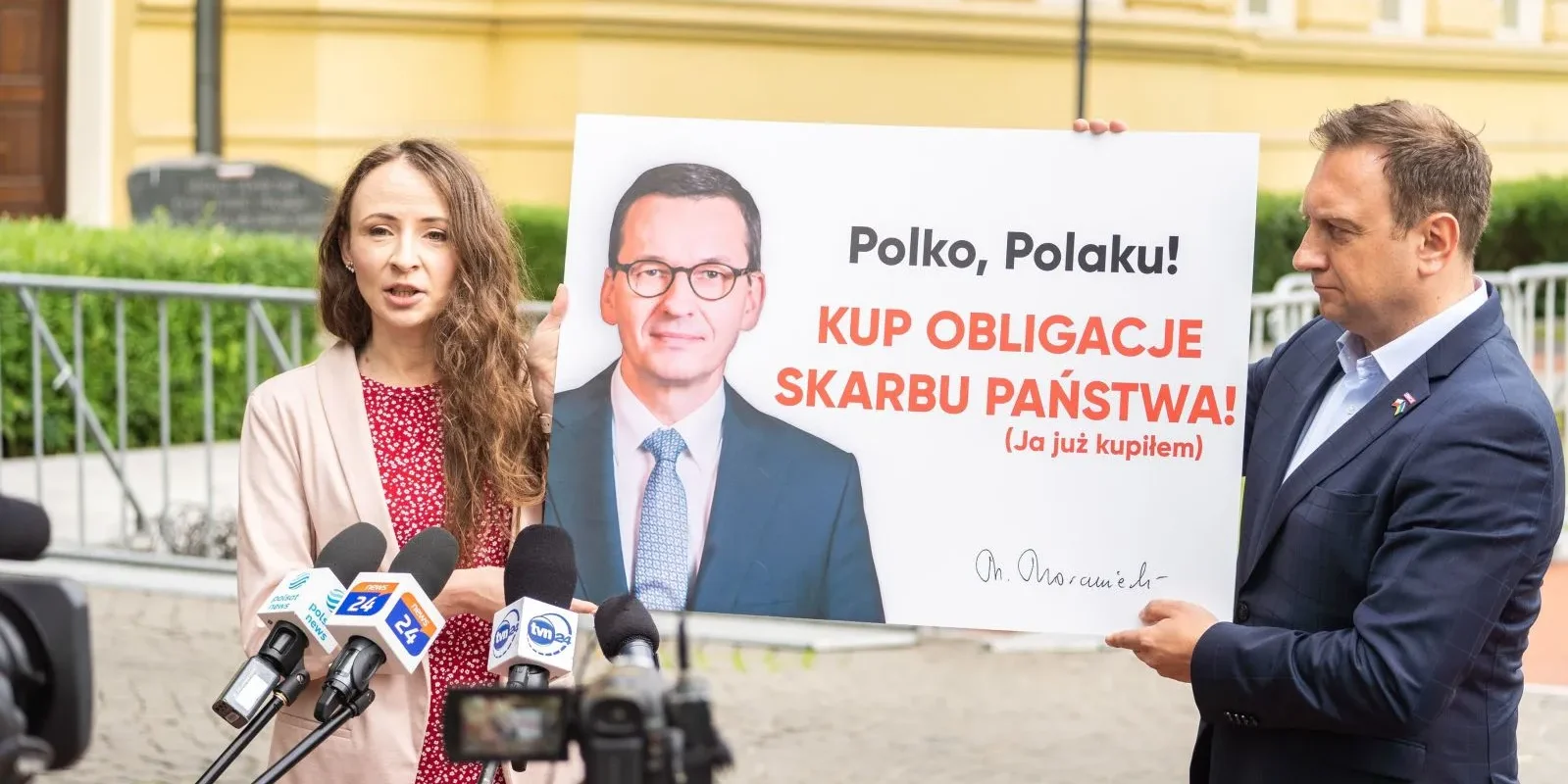 Na zdjęciu kobieta podczas konferencji prasowej trzyma plakat z wezwaniem do zakupu obligacji Skarbu Państwa, obok niej stoi mężczyzna.