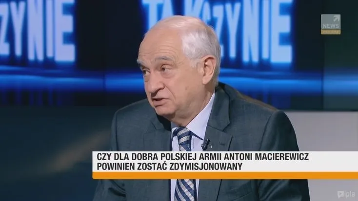 Starszy mężczyzna w garniturze wypowiada się w telewizyjnym studiu, a na pasku informacyjnym widnieje pytanie o dymisję Antoniego Macierewicza.