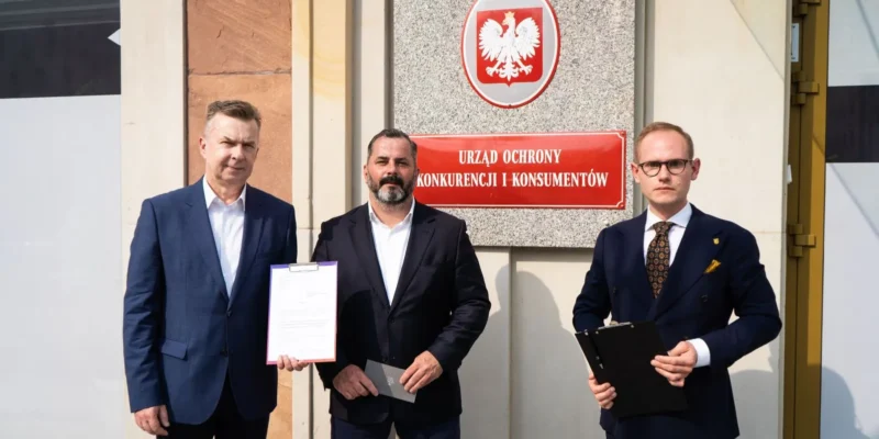 Trzej mężczyźni stoją przed budynkiem Urzędu Ochrony Konkurencji i Konsumentów, trzymając dokumenty oraz notesy.