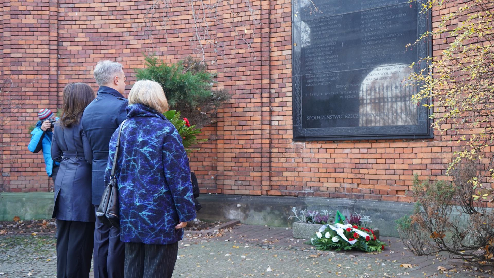 Na zdjęciu widoczna jest grupa ludzi stojąca przed tablicą pamiątkową, w milczeniu oddająca hołd złożonym kwiatom.