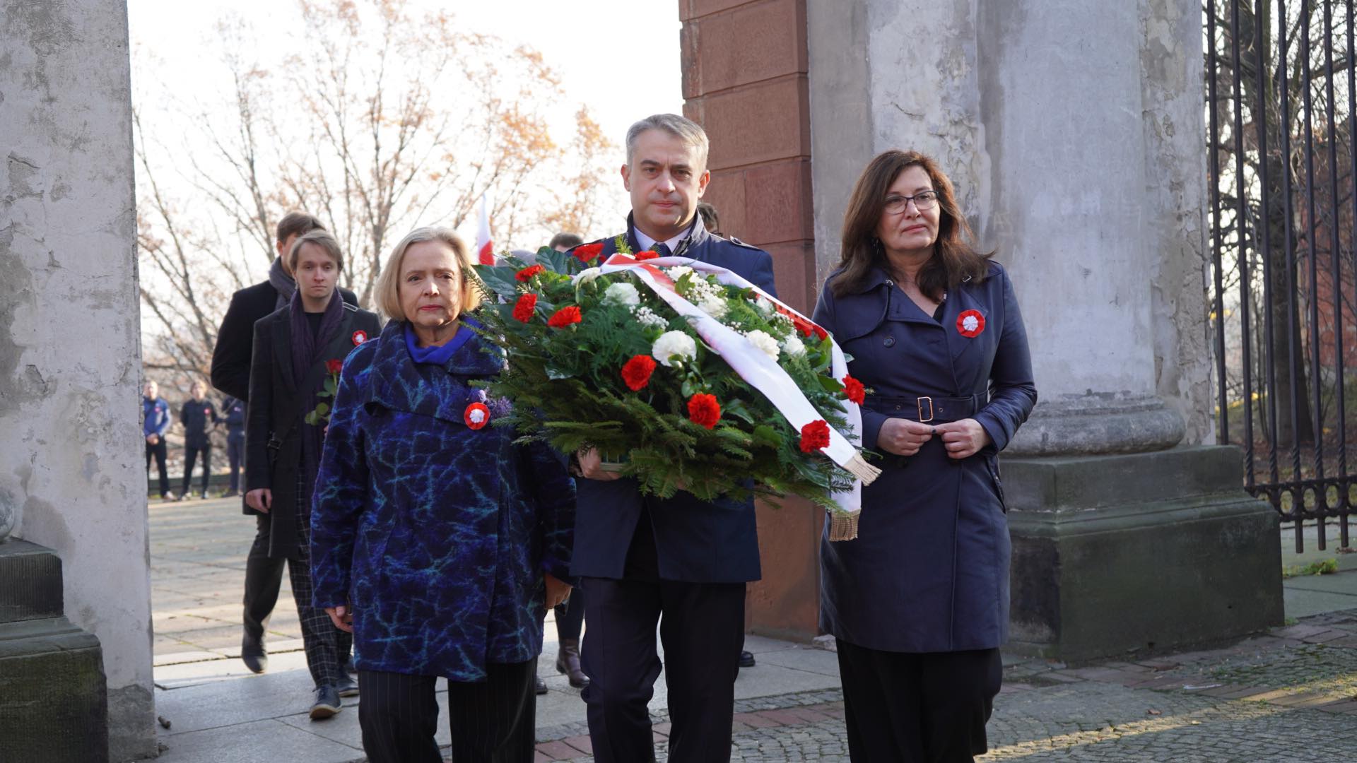 Grupa osób, w tym mężczyzna niosący wieniec z kwiatów, przechodzi przez bramę w trakcie uroczystości upamiętniającej.