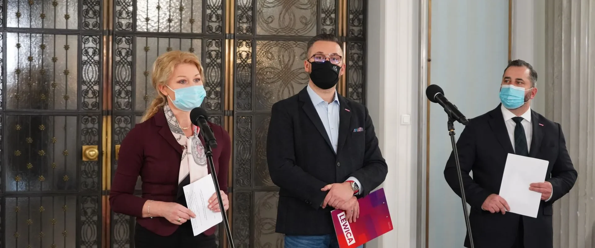 Trzy osoby w maseczkach stoją przy mikrofonach podczas konferencji prasowej w eleganckim wnętrzu z ozdobnymi drzwiami w tle, każda trzyma materiały.