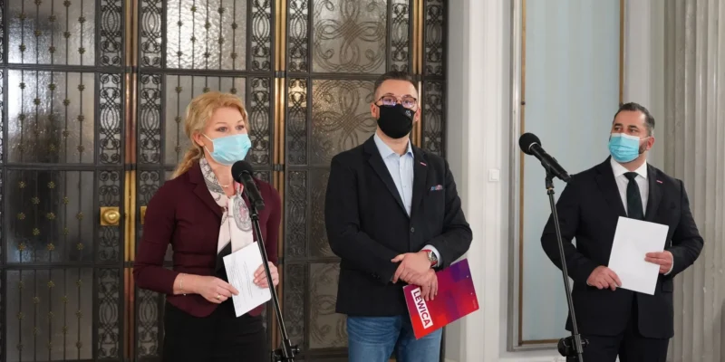 Trzy osoby w maseczkach stoją przy mikrofonach podczas konferencji prasowej w eleganckim wnętrzu z ozdobnymi drzwiami w tle, każda trzyma materiały.