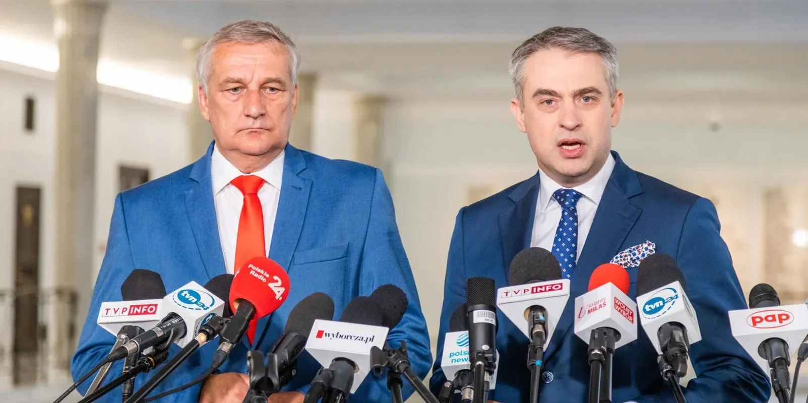 Dwaj mężczyźni w garniturach stoją przed mikrofonami na konferencji prasowej w jasnym wnętrzu.