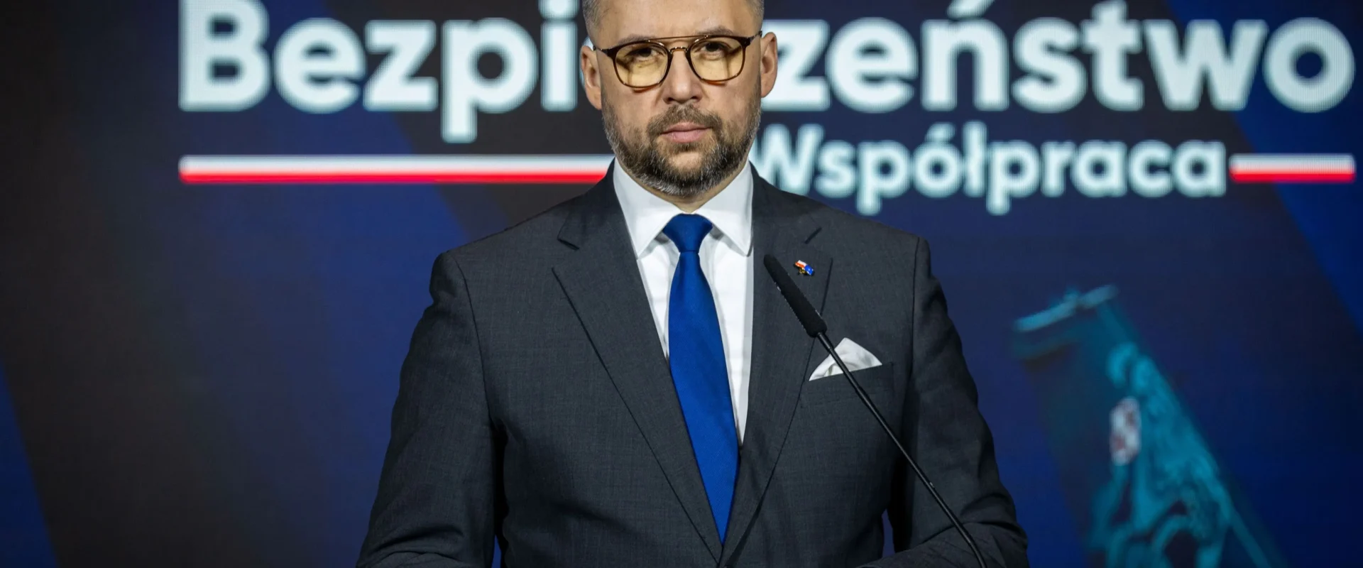 Mężczyzna w garniturze i niebieskim krawacie stoi przy mównicy podczas konferencji, a w tle widnieje duży napis „Bezpieczeństwo Współpraca”.