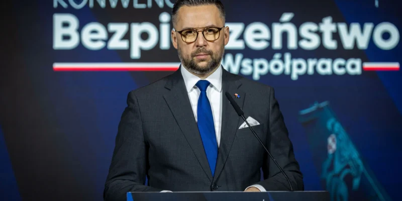 Mężczyzna w garniturze i niebieskim krawacie stoi przy mównicy podczas konferencji, a w tle widnieje duży napis „Bezpieczeństwo Współpraca”.