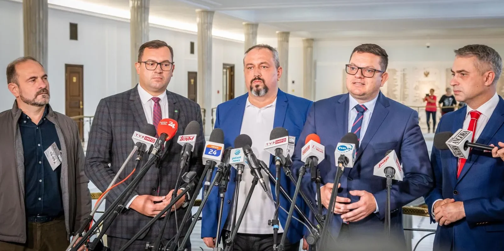 Na zdjęciu widać grupę mężczyzn ubranych w garnitury, którzy stoją przed mikrofonami i rozmawiają na konferencji prasowej.