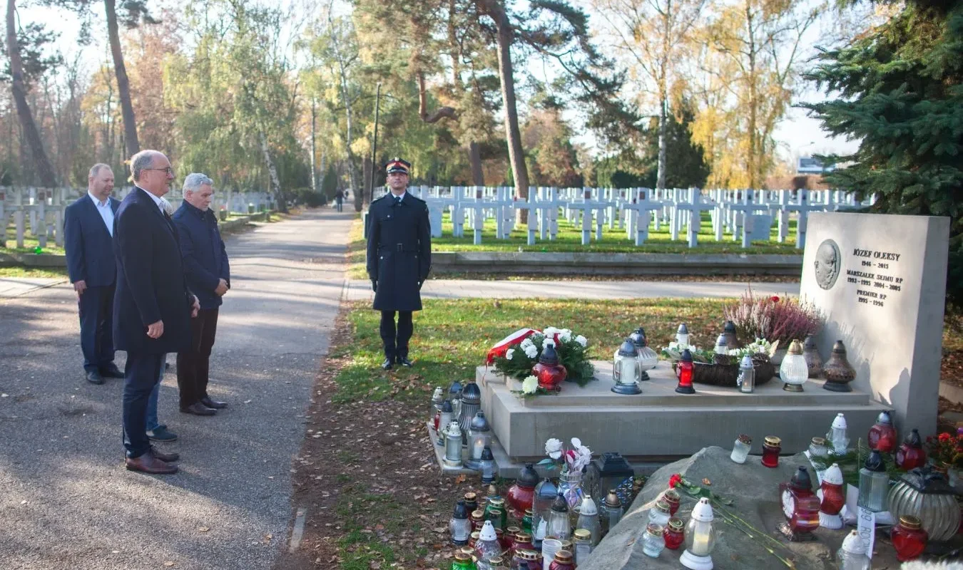 Na zdjęciu grupa osób stoi przy grobie, w tle widać cmentarz z licznymi nagrobkami, a obok grobu znajdują się znicze i kwiaty.