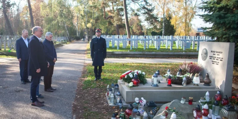 Na zdjęciu grupa osób stoi przy grobie, w tle widać cmentarz z licznymi nagrobkami, a obok grobu znajdują się znicze i kwiaty.