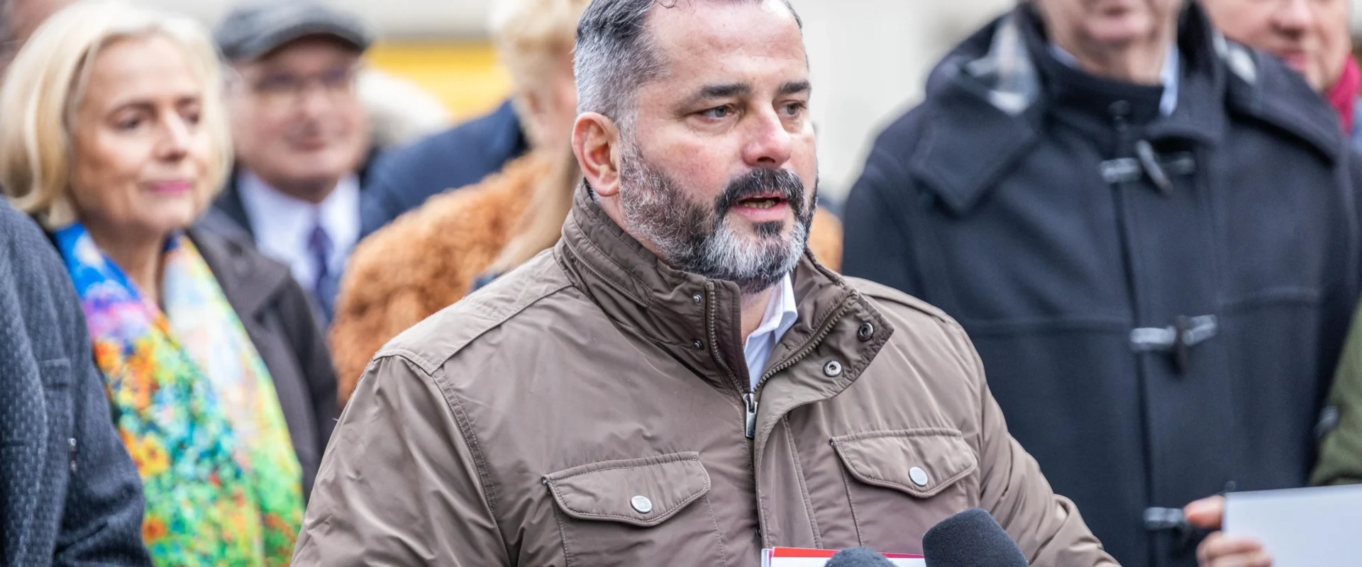 Mężczyzna w brązowej kurtce przemawia do reporterów podczas konferencji prasowej, trzymając dokumenty i stojąc przed mikrofonami.