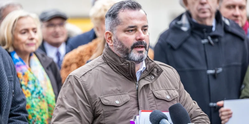 Mężczyzna w brązowej kurtce przemawia do reporterów podczas konferencji prasowej, trzymając dokumenty i stojąc przed mikrofonami.