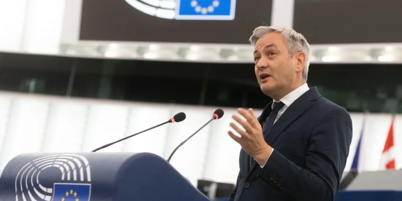 Polski polityk w garniturze przemawia z mównicy w sali plenarnej Parlamentu Europejskiego, na tle symbolu Unii Europejskiej.