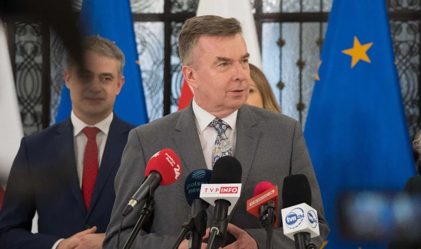 Mężczyzna w szarym garniturze przemawia na konferencji prasowej, stojąc przy mikrofonach, w tle widoczne flagi Polski i Unii Europejskiej.