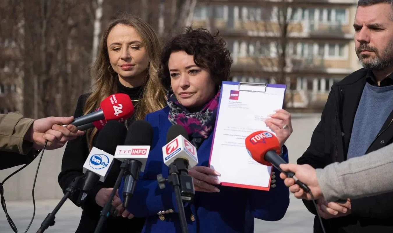 Dwie kobiety stoją na zewnątrz przed mikrofonami mediów i prezentują dokument podczas konferencji prasowej w plenerze.