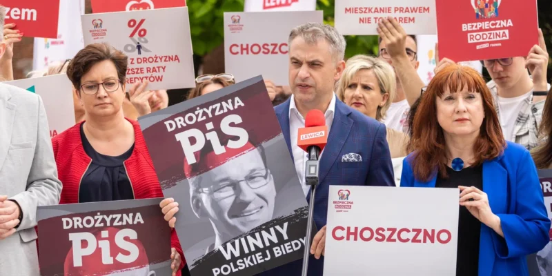 Grupa zwolenników Lewicy podczas wiecu trzyma plakaty anty-PiS i słucha przemówienia wygłaszanego do mikrofonu z logiem Lewicy.