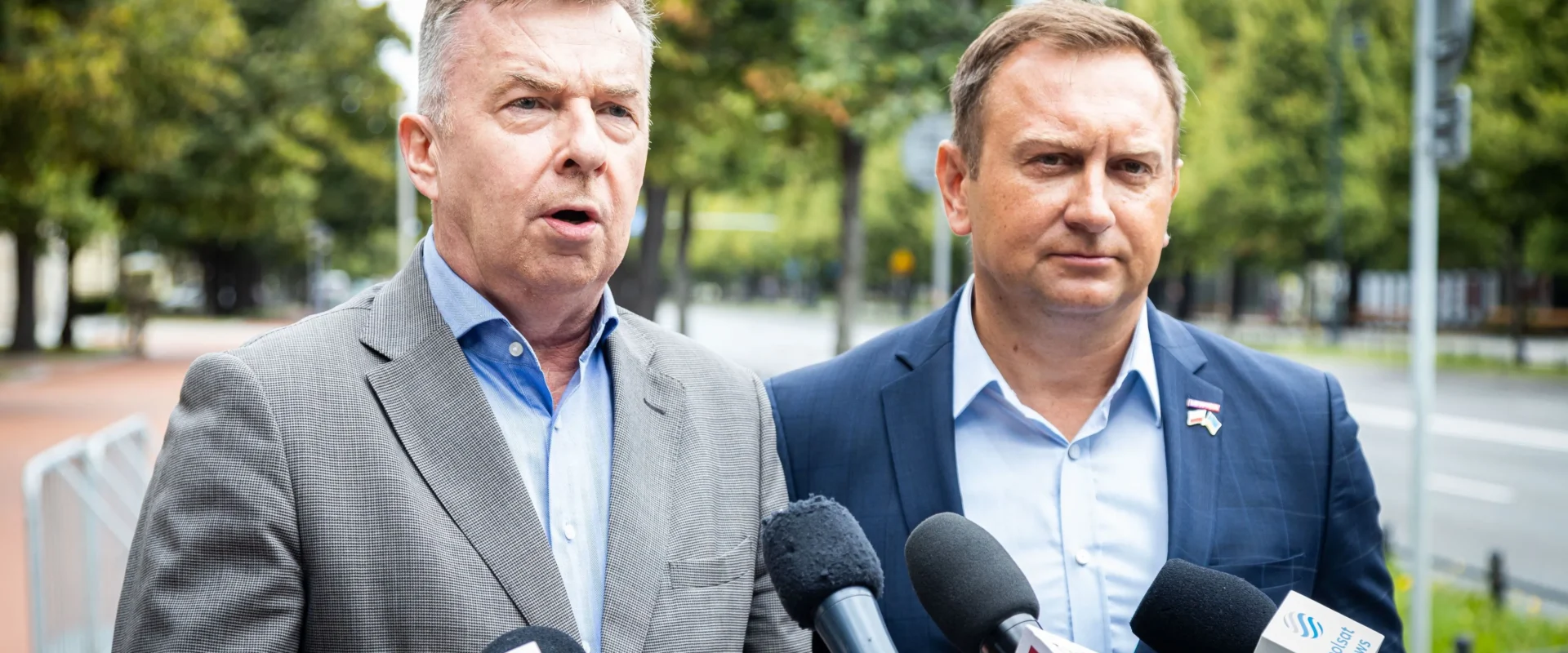 Dwaj mężczyźni w garniturach przemawiają do dziennikarzy podczas konferencji prasowej w parku.