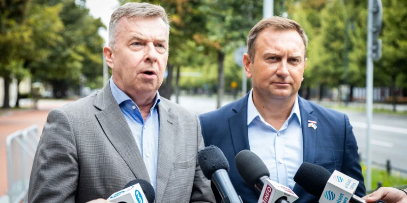 Dwaj mężczyźni w garniturach przemawiają do dziennikarzy podczas konferencji prasowej w parku.