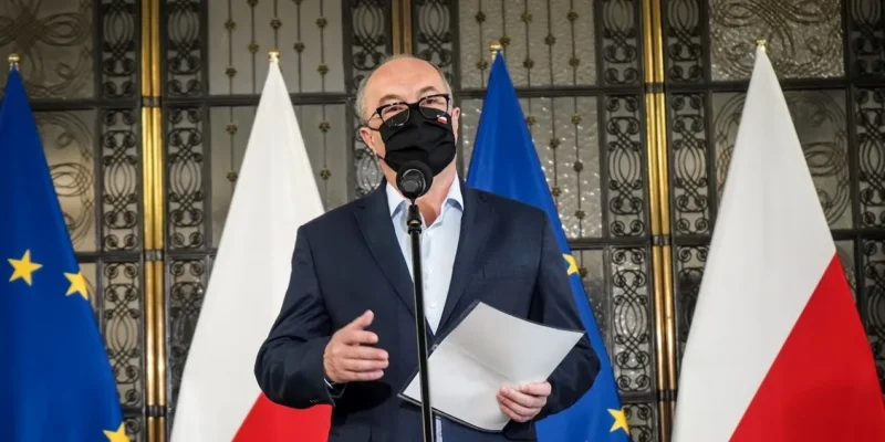 Mężczyzna w garniturze z czarną maseczką przemawia do mikrofonu, trzyma w dłoni kartkę, a w tle stoją flagi Unii Europejskiej i Polski.