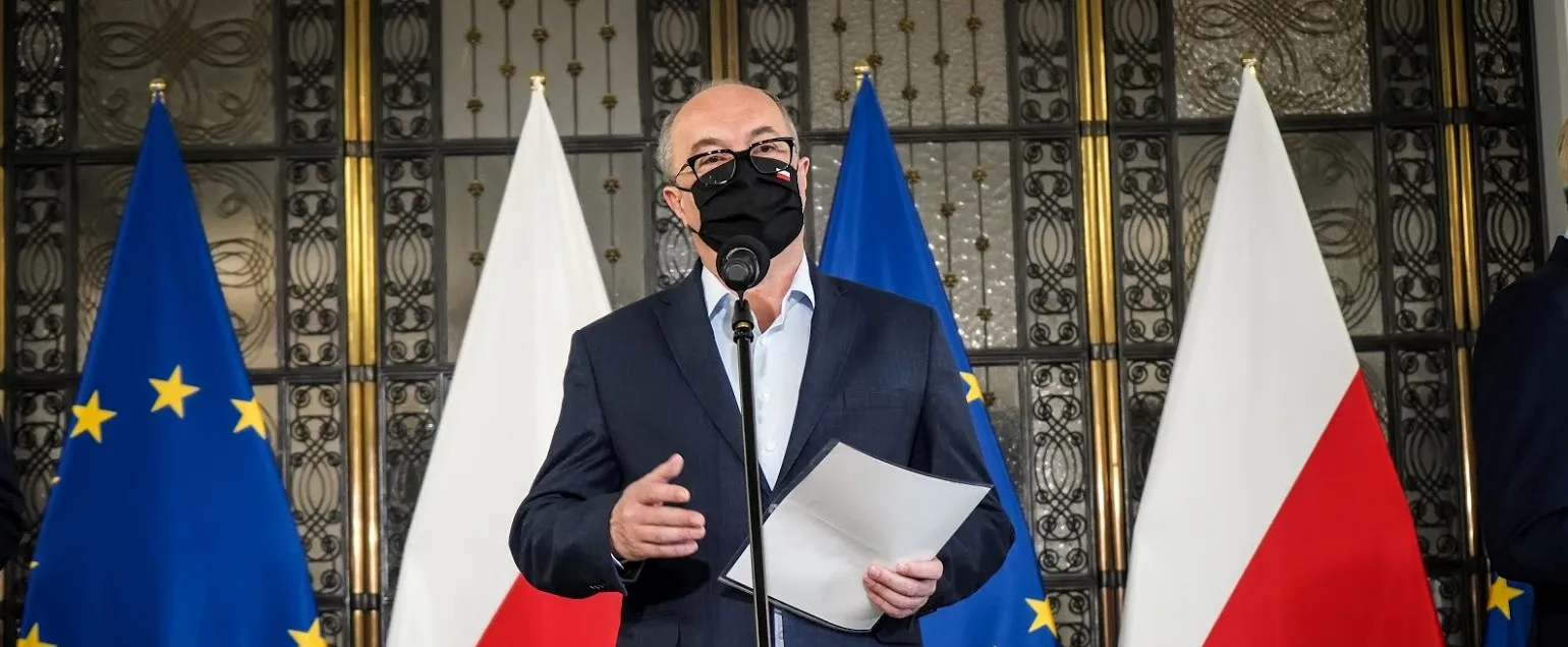 Mężczyzna w garniturze z czarną maseczką przemawia do mikrofonu, trzyma w dłoni kartkę, a w tle stoją flagi Unii Europejskiej i Polski.